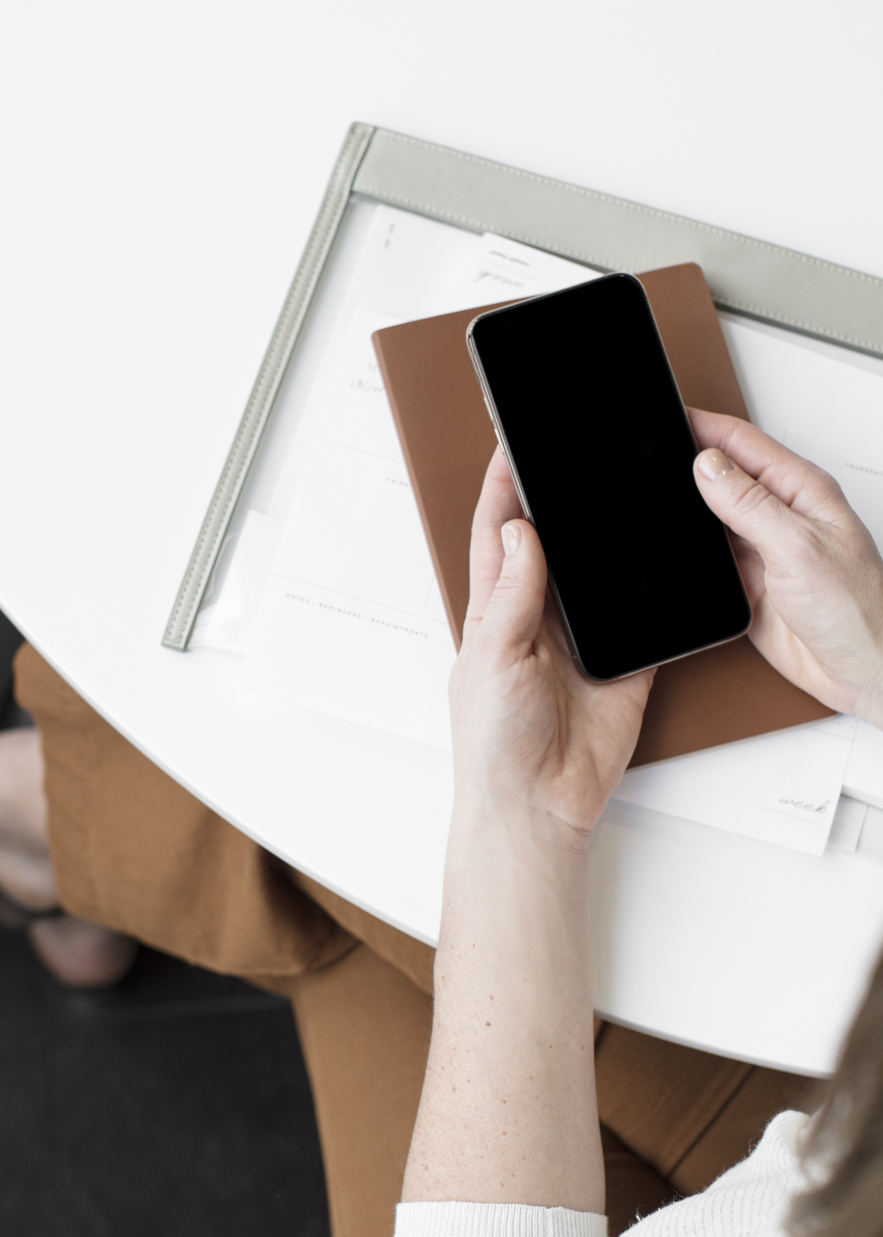 Person holding a black smartphone above a brown planner and papers on a light desk