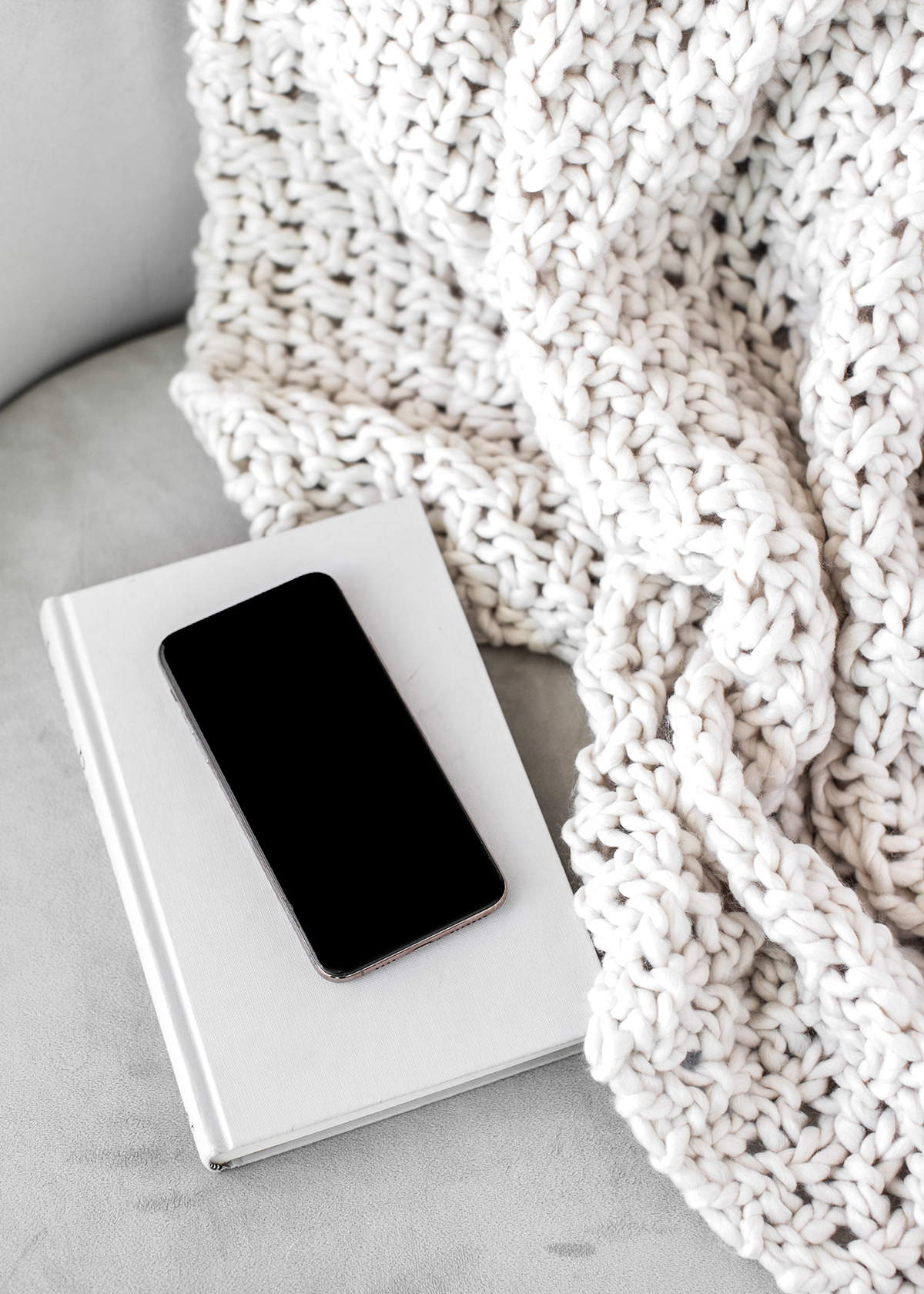 White chunky knit blanket draped over an off-white surface with a smartphone resting on a white book