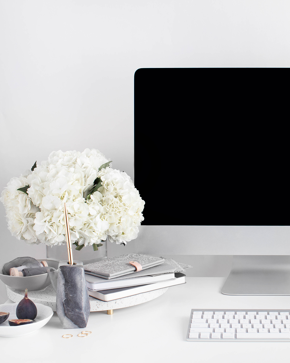 Modern desk scene with white hydrangeas, stacked books, a grey keyboard, and a black monitor